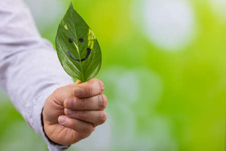 businessman holding a green leaf in his hands with a painted smile, natural background with blurred greenery, creative, work, business and personal development concept in harmony with nature, copy spaceの写真素材