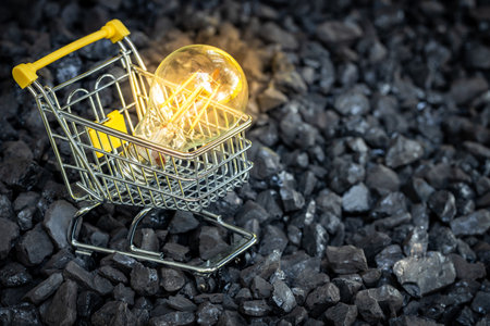 Shopping cart with a glowing light bulb standing buried in a coal heap, high costs of obtaining energy from coal, business and environmental conceptの写真素材
