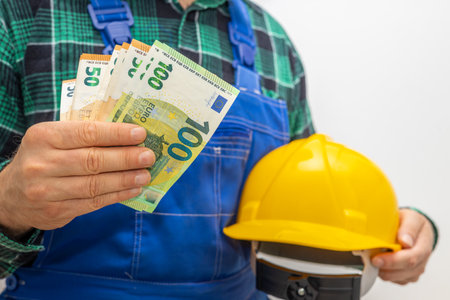 Wages in the euro area for manual workers, builders and specialists in finishing works, a specialist with a construction helmet holding a file of money in his handの写真素材