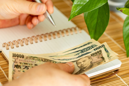 A Japanese man holding money in his hand and writing down his income and expenses in a notebook. A...