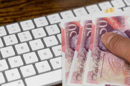 Hand holding British Pounds over white computer keyboard. Financial concept. Online shopping. UK money, GBP currency tradingの写真素材