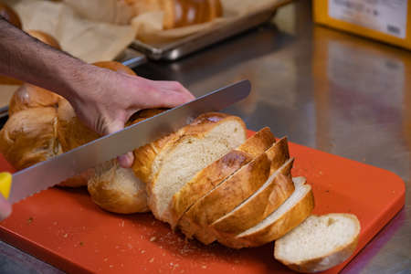 Man cutting slicing a loft of fresh swiss zopf braided bread with a serrated knife on a red cutting...