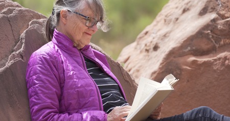 Mature outdoors enthusiast smiling and reading book in nature seated on rockの写真素材