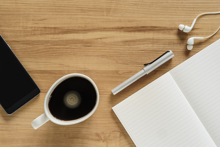 Top view of wooden table with black coffe, blank note, pen, mobile phone and earphoneの写真素材