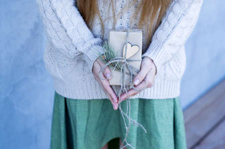 Time gifts - gift box in hand girls. Christmas or new year gift. Girl in a knitted sweater holds gift box in brown craft paper wih heart. Christmas, hew year, birthday concept.の写真素材