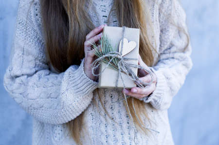 Time gifts - gift box in hand girls. Christmas or new year gift. Girl in a knitted sweater holds gift box in brown craft paper wih heart. Christmas, hew year, birthday concept.の写真素材