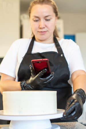 Woman blogger takes photos of cakes for her followers. Pastry chef cook confectioner or baker photographing fresh cake on cake stand in kitchen at home. Concept of homemade pastry, hobby.の写真素材