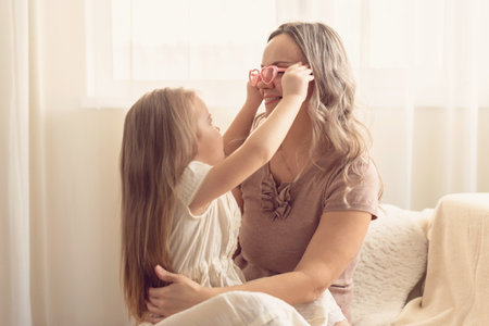 Smiling preschool girl sitting on mommies lap at home. Carefree mom and little girl laughing while playing at home. Love, warm family relationships. Happy kid with mum spending free time togetherの写真素材