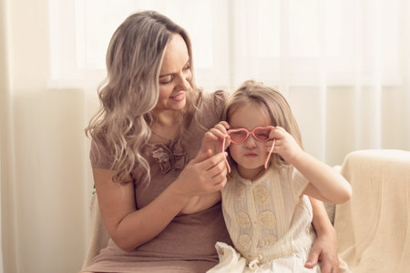 Smiling preschool girl sitting on mommies lap at home. Carefree mom and little girl laughing while playing at home. Love, warm family relationships. Happy kid with mum spending free time togetherの写真素材