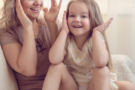 Smiling preschool girl sitting on mommies lap at home. Carefree mom and little girl laughing while playing at home. Love, warm family relationships. Happy kid with mum spending free time togetherの写真素材
