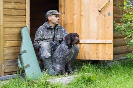 hunter and his dog sitting at the door of a wooden house, next to a shotgun in a caseの写真素材
