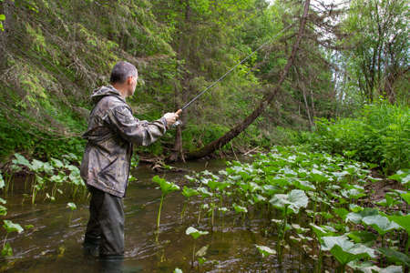 fisherman with a spin fishing rod in his hands performs retrieval lure on a narrow forest riverの写真素材