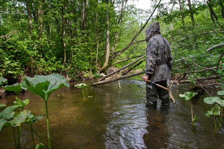 a fisherman with a spinning rod in his hand stands in front of a blockage on a forest riverの写真素材