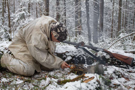 a hunter dressed in white camouflage builds a fire using matches in the winter forestの写真素材