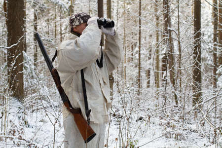 a hunter, dressed in white camouflage, with a shotgun on his shoulder, watches the winter forest through binocularsの写真素材