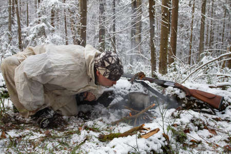 a hunter dressed in white camouflage blows a fire in the winter forestの写真素材