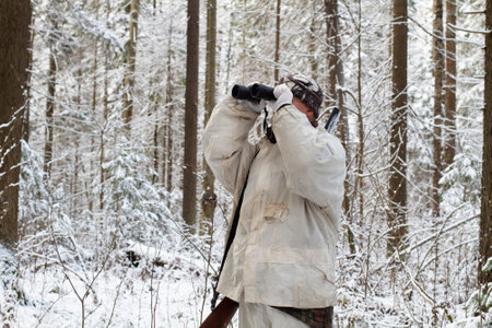 The hunter, dressed in white camouflage, with a shotgun on his shoulder, looks through binoculars. Around the snow-covered northern forest. Medium long shot.の写真素材