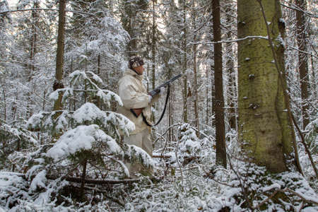 The short winter day was ending, and twilight had begun in the snow-covered forest. A hunter dressed in a white camouflage suit stands among the trees. He is alert and holds a shotgun in his hands.の写真素材
