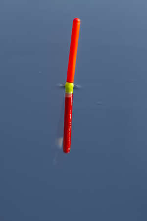 the orange tip of the fishing float sits vertically in the smooth blue water of the lake, close upの写真素材