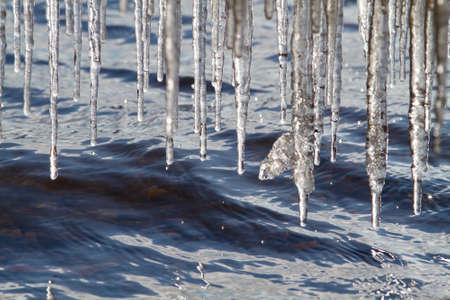 It is a sunny spring day. Many jagged icicles hang over the choppy blue water. Drops fall from them.の写真素材