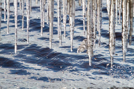 It is a sunny spring day. Many icicles hang over the choppy blue water. Numerous drops fall from them.の写真素材