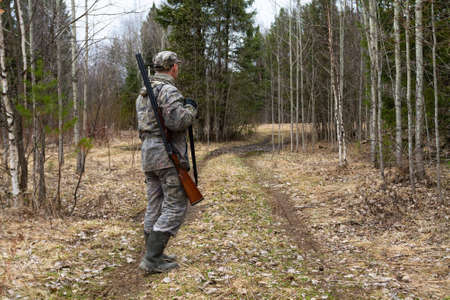 A hunter walks along a road in the spring forest. He has a shotgun slung over his shoulder. The evening twilight deepens.の写真素材