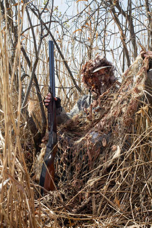 The duck hunter disguises himself in the bushes using a camouflage net. He holds a shotgun in his hand.の写真素材