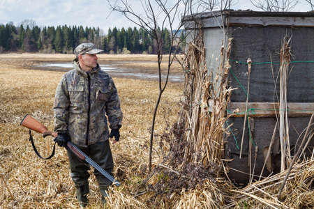 A duck hunter stands next to an old hunting shelter. He has a shotgun in his hands. Behind the hunter, the muddy shallow water begins, where the ducks arrive.の写真素材