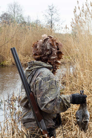 The hunter is standing on the shore of a lake that is overgrown with reeds. He holds in his hands his prey - two shot ducks. He has a shotgun slung over his shoulder.の写真素材
