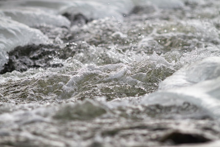 A fast river stream flows among the ice. Its surface is covered with foam and splashes. Selective focusing.の写真素材