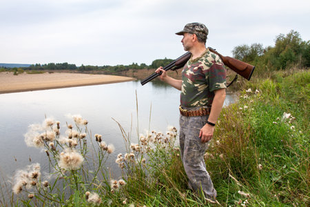 The hunter stands on the bank and looks at the river. He carries an unloaded shotgun on his shoulder.の写真素材