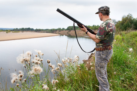 The hunter is standing on the bank of the river, in summer. He is holding a shotgun in his hands.の写真素材