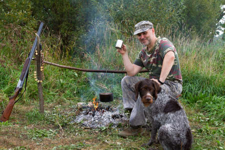 A hunter and his dog are in a summer field camp. A pot hangs over the campfire. The hunter drinks tea from a mug. A shotgun and a bandolier hang nearby.の写真素材
