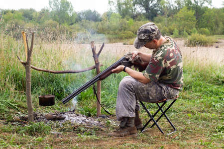 The hunter is sitting on a folding chair next to the campfire. A pot hangs over the fire. The hunter wipes the shotgun with a rag.の写真素材