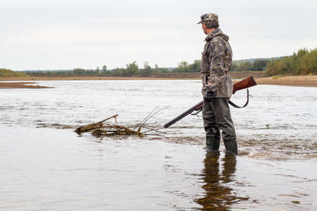 A hunter with a shotgun in his hand is standing in shallow water. He wades across the river.の写真素材
