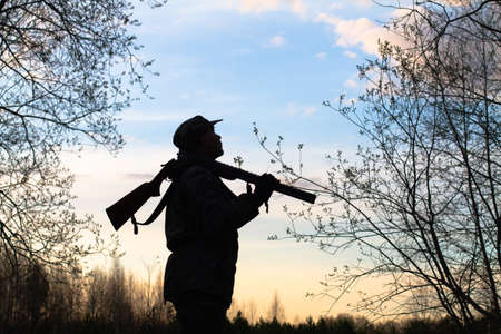 The black silhouette of a hunter with an unloaded shotgun on his shoulder stands against the background of the evening sunset. He looks at the sky.の写真素材