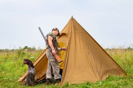 A hunter stands at the entrance to a hunting tent. A hunting dog is sitting nearby. Around there is a field overgrown with grass.の写真素材