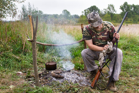 A hunter is sitting on a folding chair in a field camp. A pot hangs over the campfire. The hunter wipes the shotgun with a rag.の写真素材