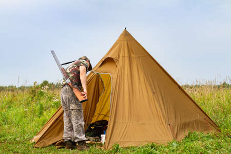 The hunter is in a hunting camp. He looks inside the tent. Around there is a field overgrown with grass.の写真素材