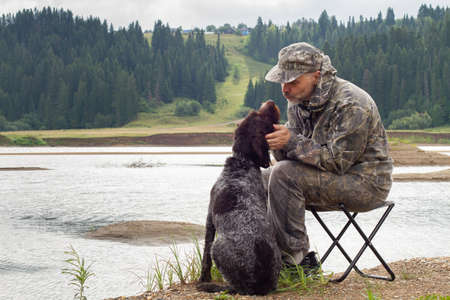 The river bank. The hunter is sitting on a folding chair. His dog is sitting next to him.の写真素材