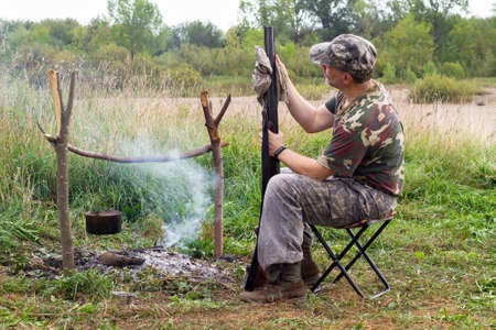The hunter wipes the gun with a rag. He is sitting on a folding chair next to the campfire. A pot hangs over the fire.の写真素材