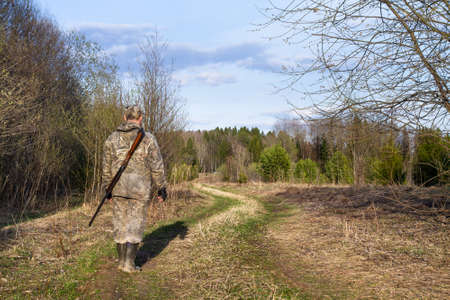 A hunter walks along the road in the spring forest. He's got a shotgun on his back. Light from the low evening sun.の写真素材