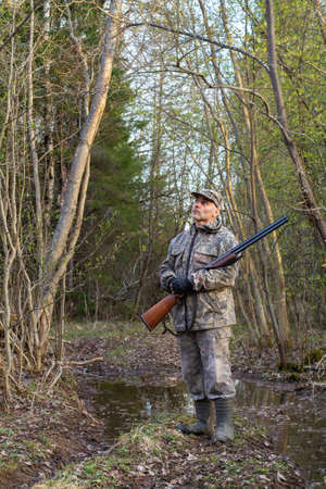 A hunter stands in a thicket of bushes in spring. Evening twilight is gathering. The hunter is holding a shotgun. He's waiting for the woodcock to arrive.の写真素材
