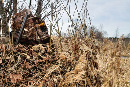 The hunter disguised himself in a shelter and lures a waterfowl with a duck call. He holds a shotgun in his hand.の写真素材