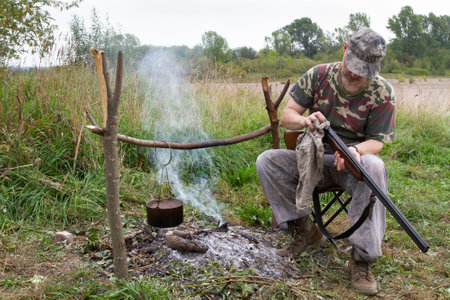 The hunter is sitting on a folding chair next to the campfire. A pot hangs over the fire. The hunter wipes the weapon with a rag.の写真素材
