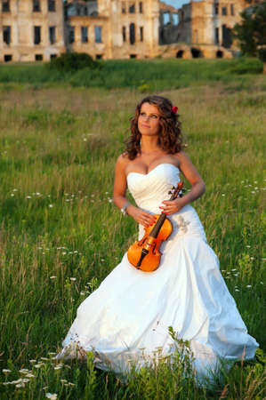 Smiling bride with violin standing in front of old ruins at sunsetの写真素材
