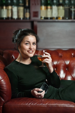 Smiling young woman with a glass of brandy lying on the sofa