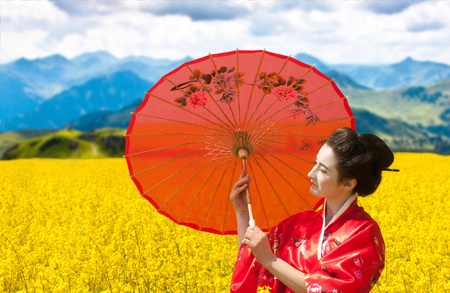 Asian style portrait of a woman with the red umbrella in the yellow flowering field, mountains backgroundの写真素材