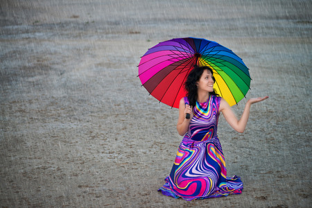 Smiling woman with colorful umbrella sitting on the ground under the rainの写真素材