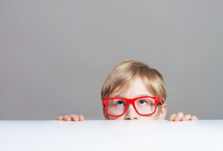 Serious boy in red-framed eyeglasses looking up from behind the tableの写真素材